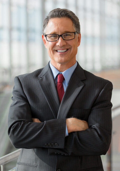 A man in a suit is smiling and standing with his arms crossed. In the background is a big open lobby with very high glass windows and the top half of an escalator.