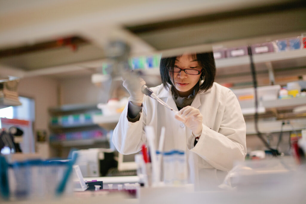 A middle-aged Asian woman with glasses is wearing a white lab coat. She's working on something in a lab.