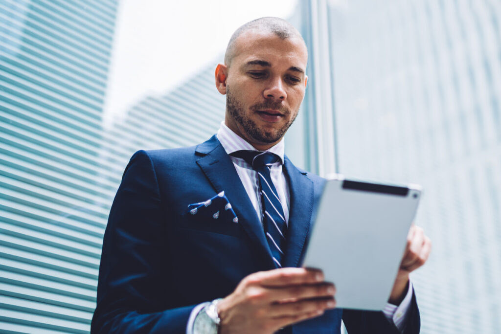 Below view of successful male executive in formal wear reading financial news on modern tablet standing on street of business city with modern skyscrapers