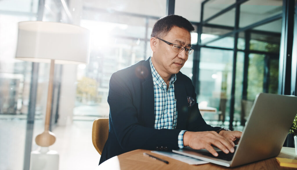 South-Asian male executive with glasses typing on a gray laptop computer. The background is well-lit.